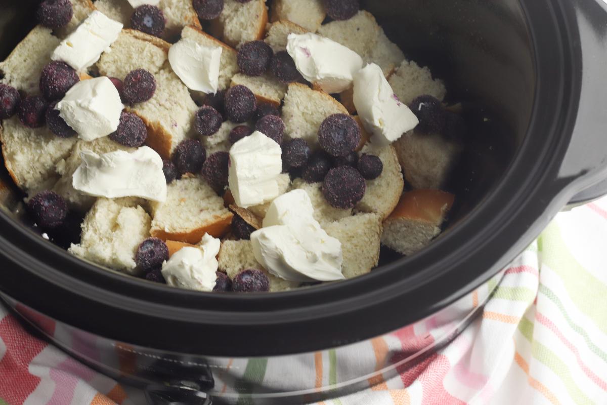 cream cheese cubes and blueberries over the top of a layer of bread cubed in the slow cooker for a delicious lazy weekend breakfast casserole.