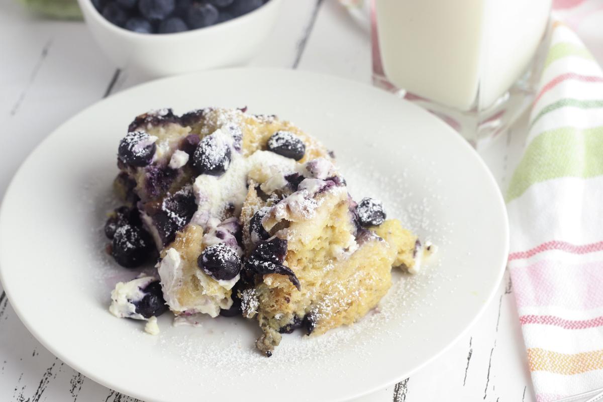 a plate with a portion of the blueberry and cream cheese french toast breakfast casserole on, behind you can just see a bowl with some blueberries in and a small milk bottle.