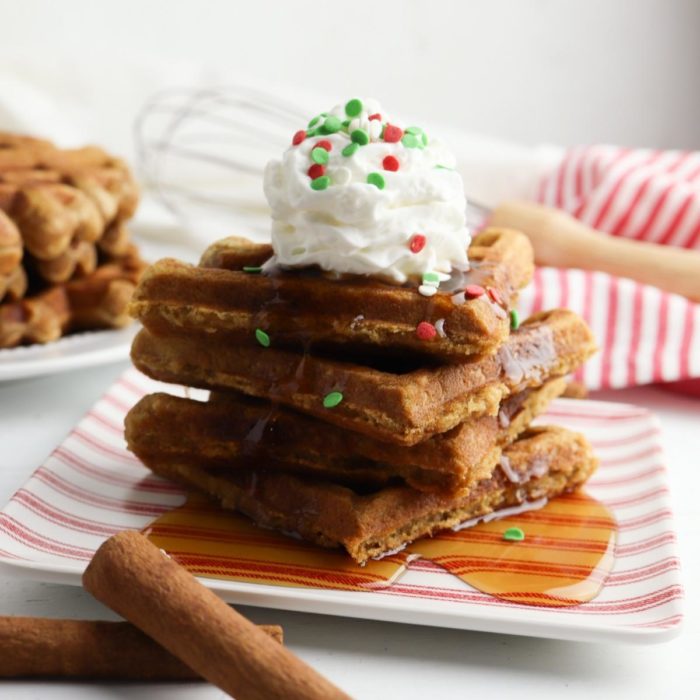 a stack of homemade gingerbread waffles on a plate with maple syrup poured over and topped with cream and christmas sprinkles