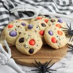 A board of delicious and simple to bake Halloween Cookies with Spooky eye balls and Halloween M&Ms presented on a chopping board with skeleton arms and fake spiders around it.