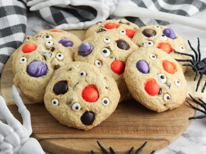 A board of delicious and simple to bake Halloween Cookies with Spooky eye balls and Halloween M&Ms presented on a chopping board with skeleton arms and fake spiders around it.