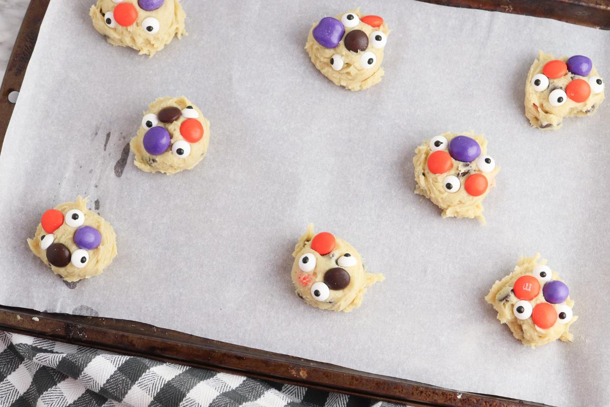 Scoops of Halloween cookie dough on a baking tray with more Halloween M&Ms added as well as candy eye balls.