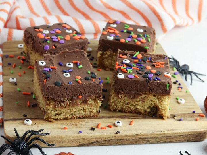 4 simple Halloween blondies with a chocolate buttercream frosting on a wooden board. The top of the cake bars is covered with Halloween sprinkles and candy eyes.