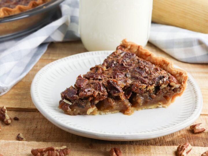 A slice of delicious homemade pecan pie on a white plate with pecan halves around the base and a glass of milk in the background.