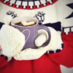 hands in mittens holding a cup of hot cocoa with a heart marshamallow in it.
